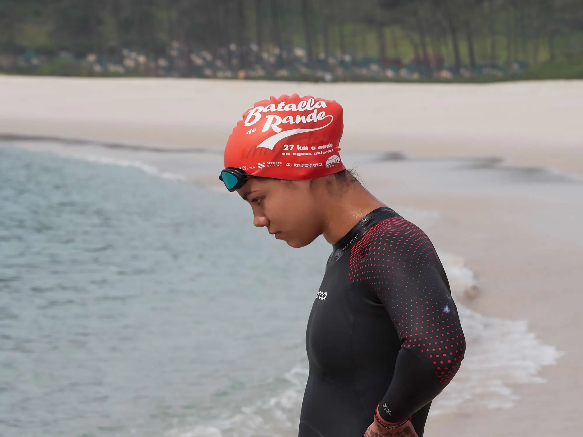 Una nadadora muy joven entra en el agua en la playa de Rodas, cabizbaja, con el gorro naranja de la Batalla de Rande que anuncia los 27 km en aguas abiertas